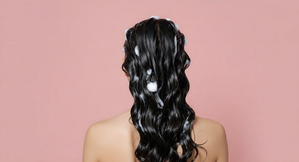 Woman with suds on wavy black hair on a pink background.