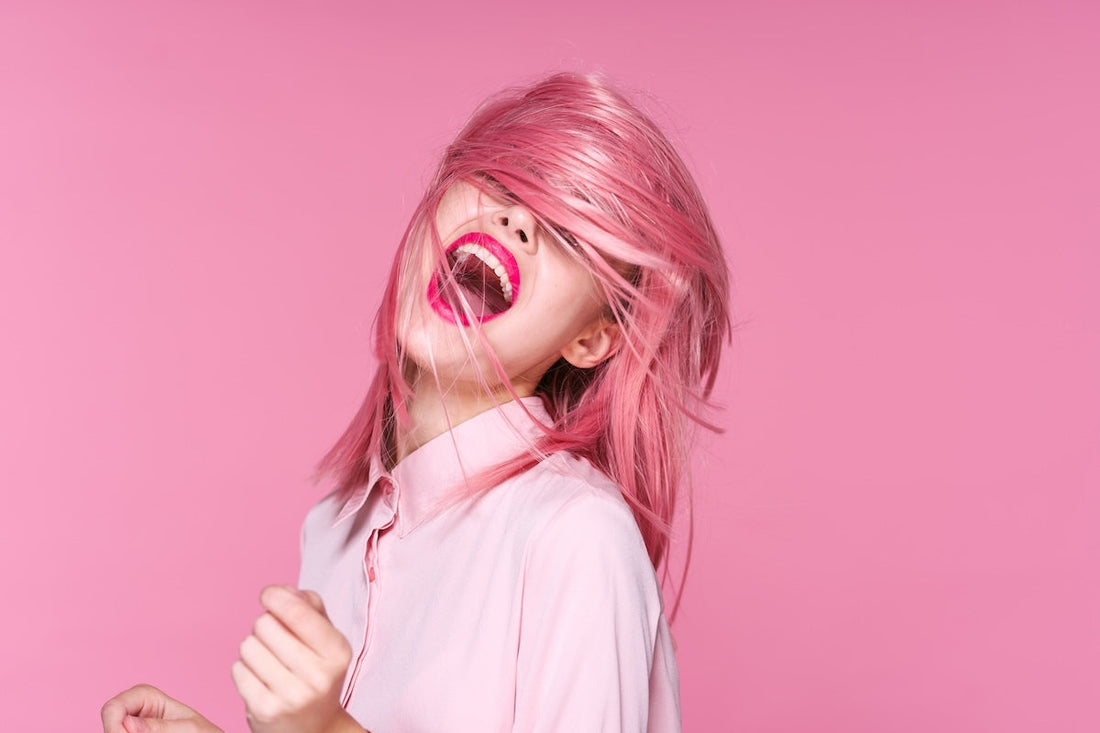 A woman with pink hair smiling in front of a pink background.