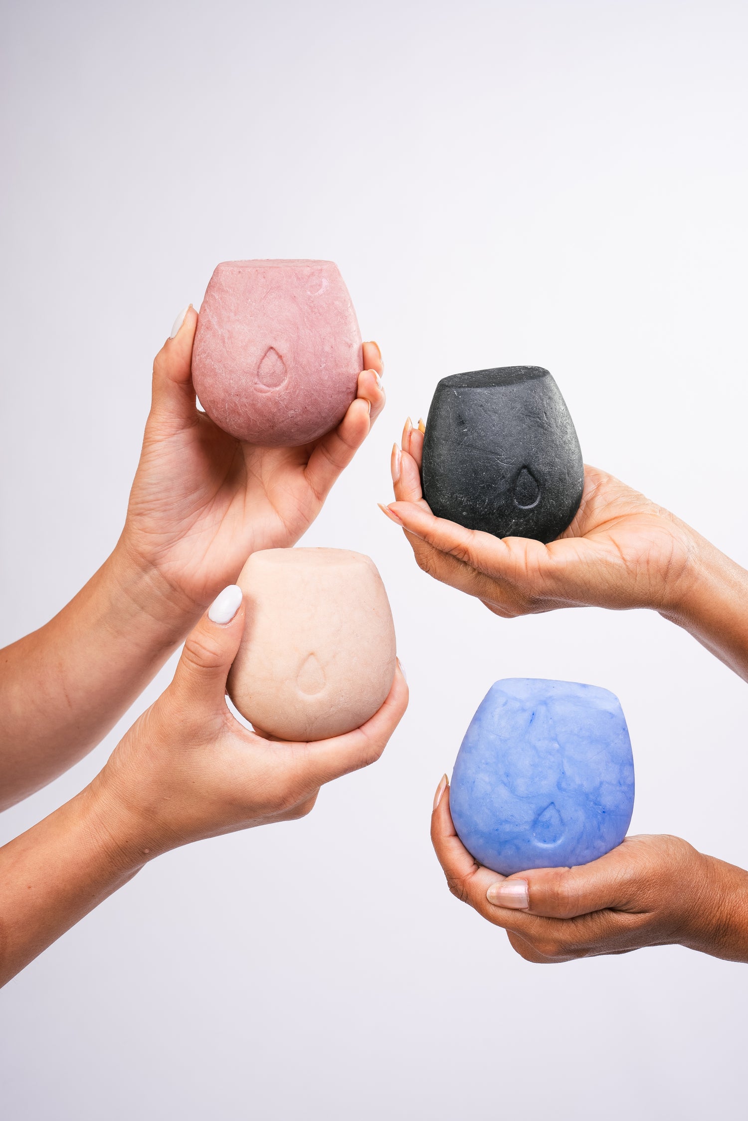 Two pairs of hands holding four body wash bars of different colors on a white background.