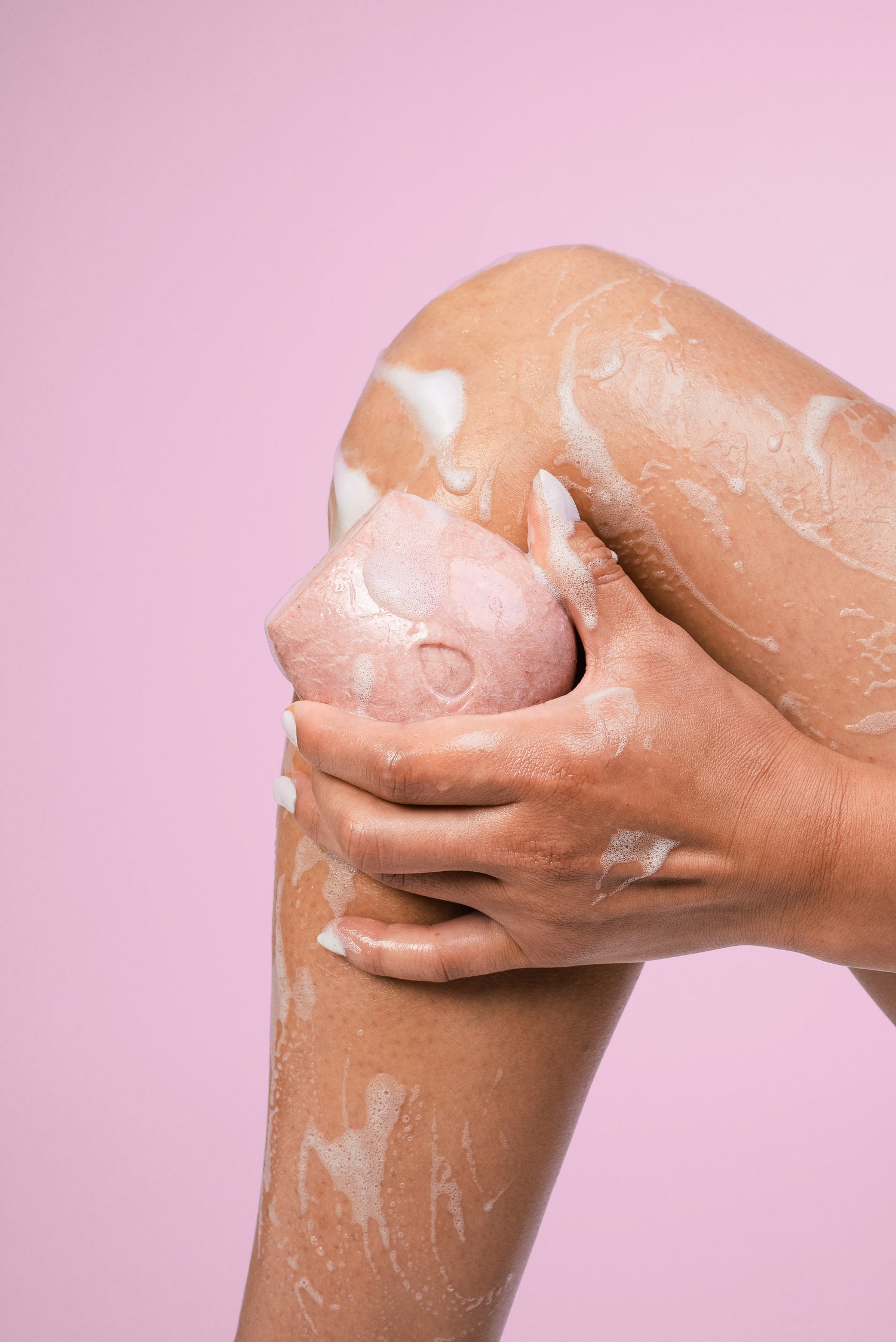 Woman using a wet pink body wash bar on her leg on a pink background.