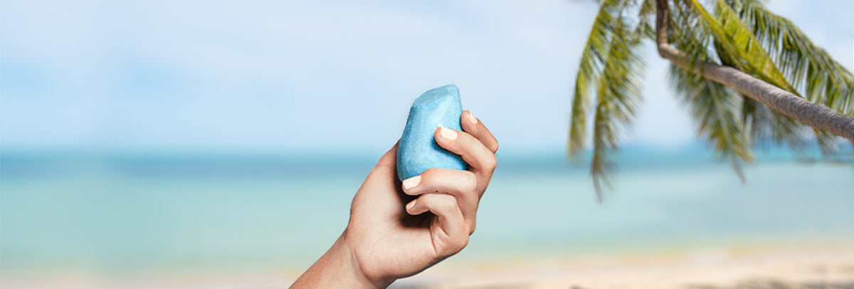 A hand holding a shampoo bar with beach in the background.