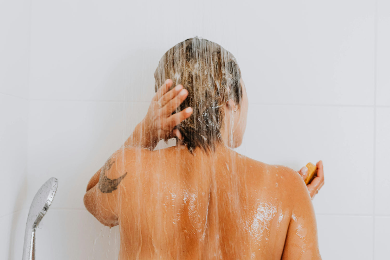 Woman in a shower washing her wet hair against a white tile background.